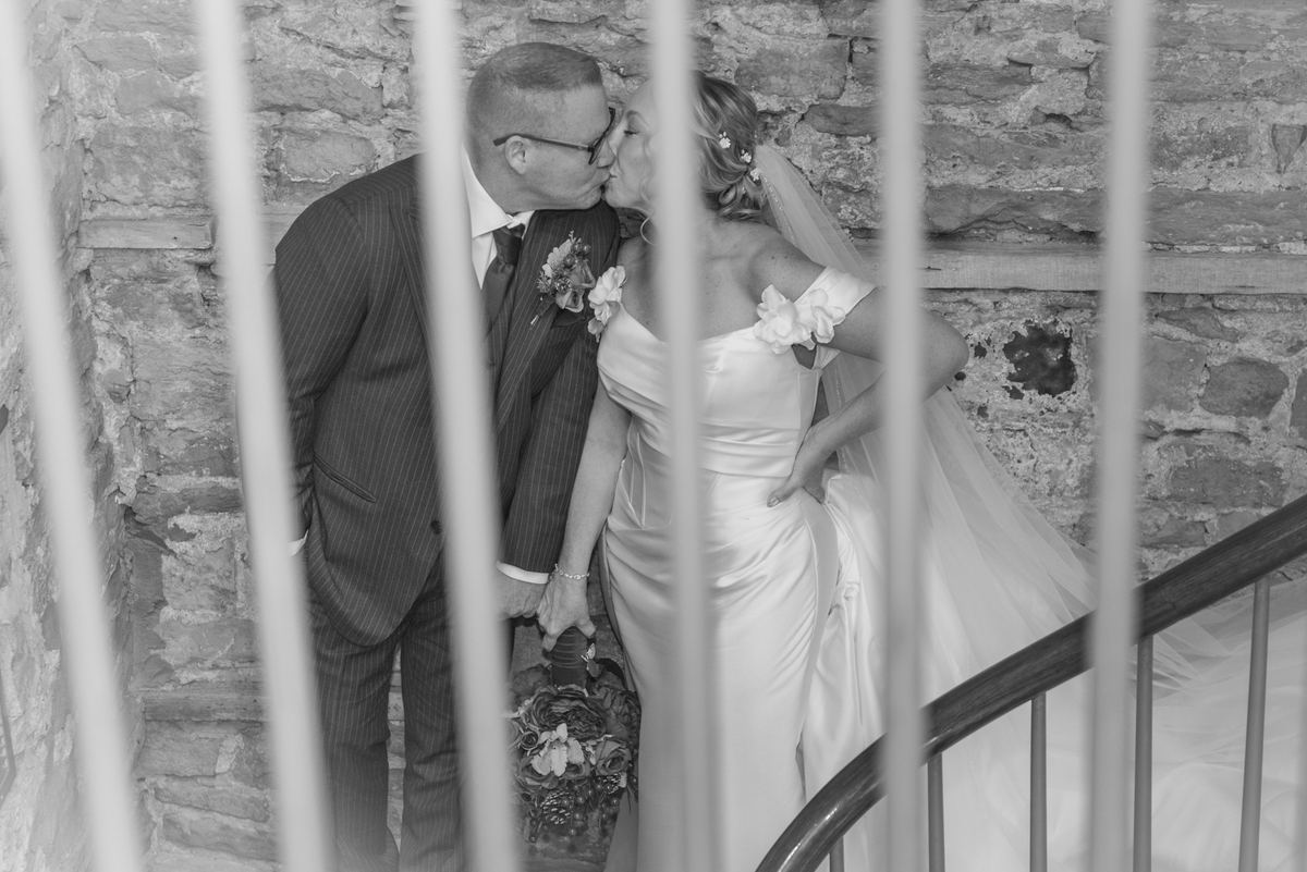 A newlywed couple kisses while climbing a stone staircase, framed by a railing, in soft light highlighting their attire.