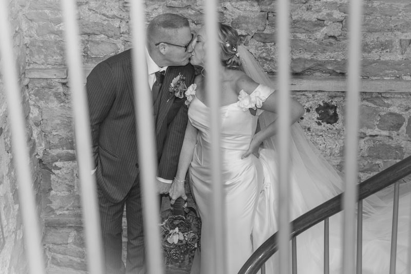 A newlywed couple kisses while climbing a stone staircase, framed by a railing, in soft light highlighting their attire.