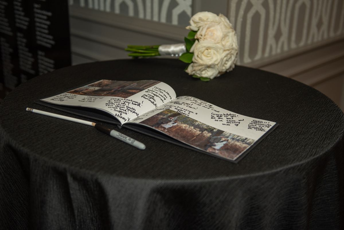 An open guest book with signatures and a white rose bouquet on a black-draped table in soft lighting.