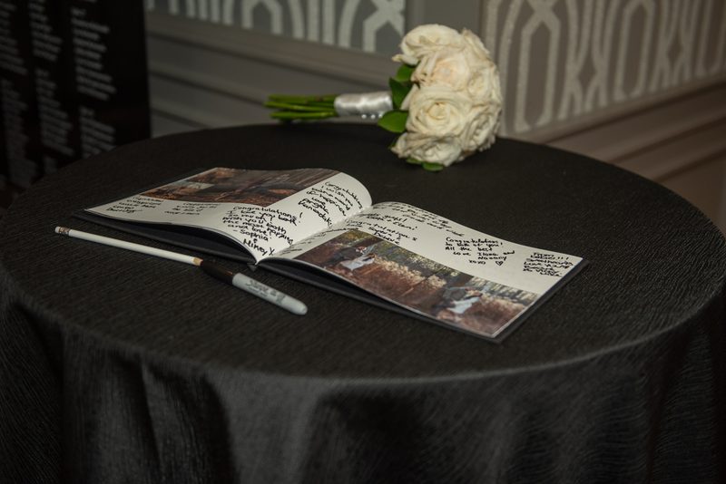 An open guest book with signatures and a white rose bouquet on a black-draped table in soft lighting.