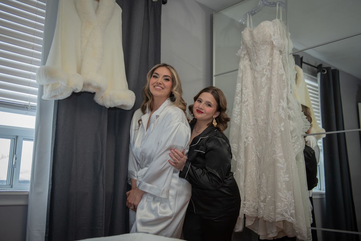 Two women smile joyfully in a bridal dressing room, surrounded by wedding attire and soft natural light.