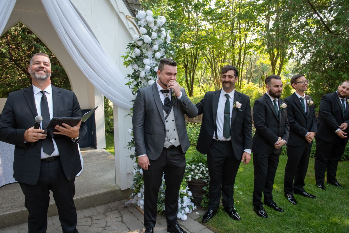 A groom addresses his guests beside a floral arch during an outdoor ceremony, sunlight filtering through trees.