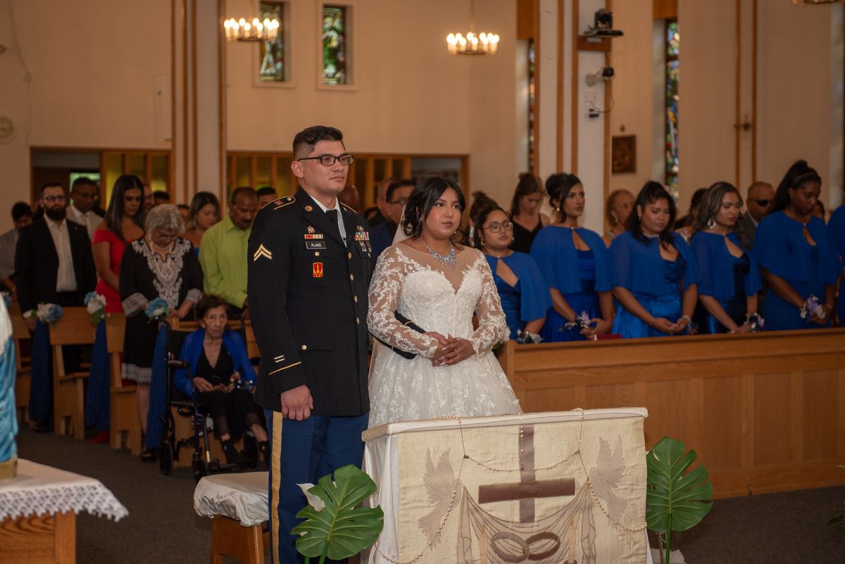 A bride and groom stand at the front of a church during a wedding ceremony, illuminated by chandeliers.