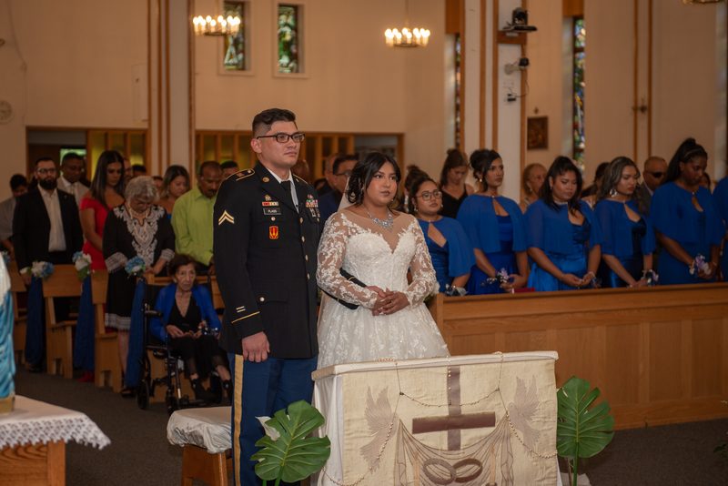 A bride and groom stand at the front of a church during a wedding ceremony, illuminated by chandeliers.