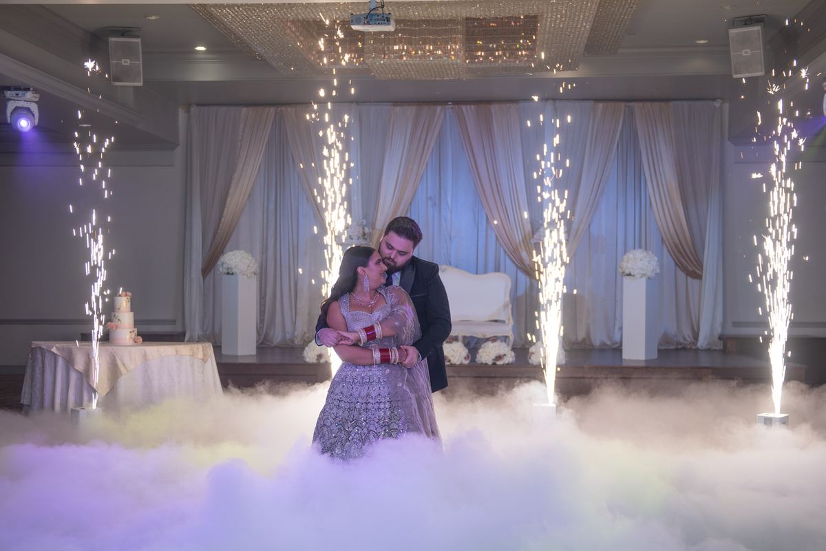 Jasleen and Marco share an intimate embrace at their grand wedding, surrounded by billowing white fog and vertical fireworks.