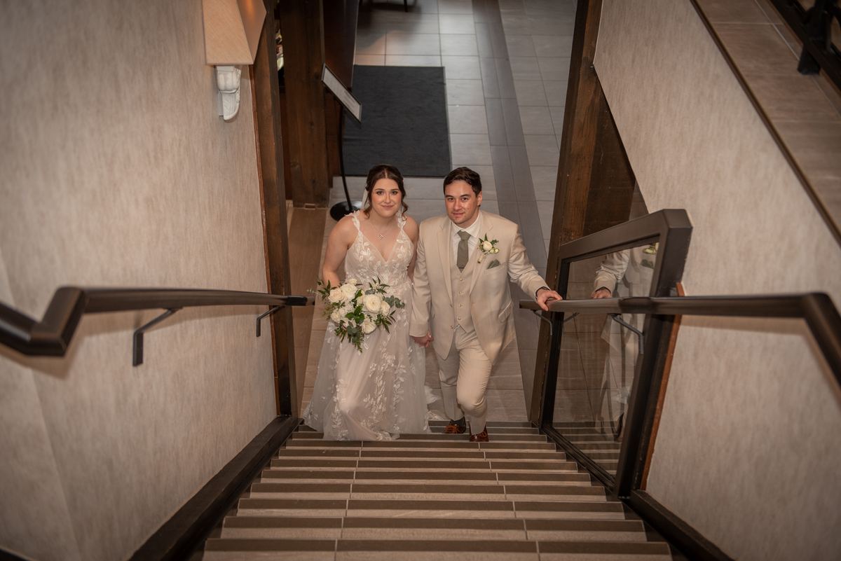 A bride and groom descend a modern staircase, smiling in their wedding attire with flowers and boutonnière.