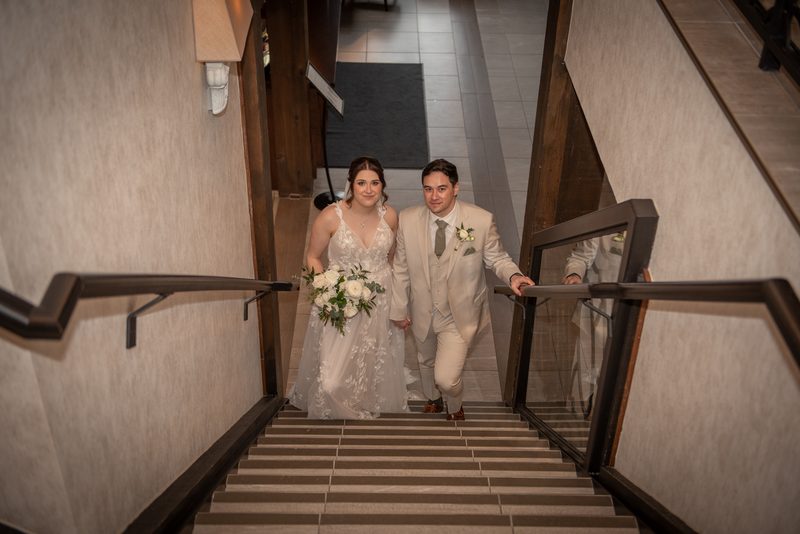 A bride and groom descend a modern staircase, smiling in their wedding attire with flowers and boutonnière.