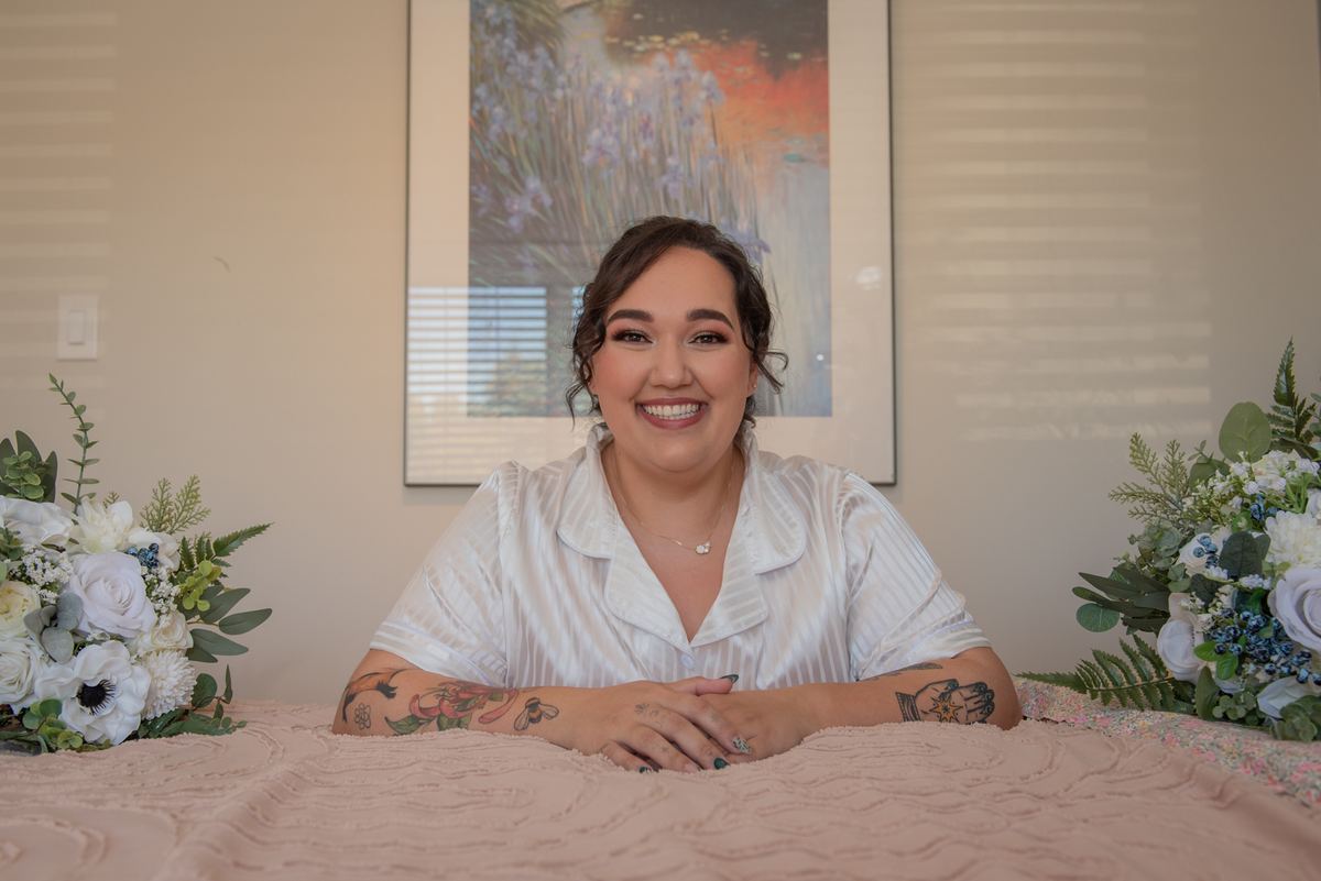 A smiling woman with tattoos sits at a pink-draped table, surrounded by floral arrangements and a painting of irises.