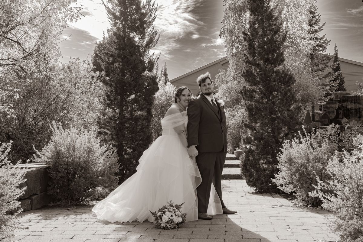 A newlywed couple in a sepia-toned infrared style, standing close on a stone pathway surrounded by lush foliage.