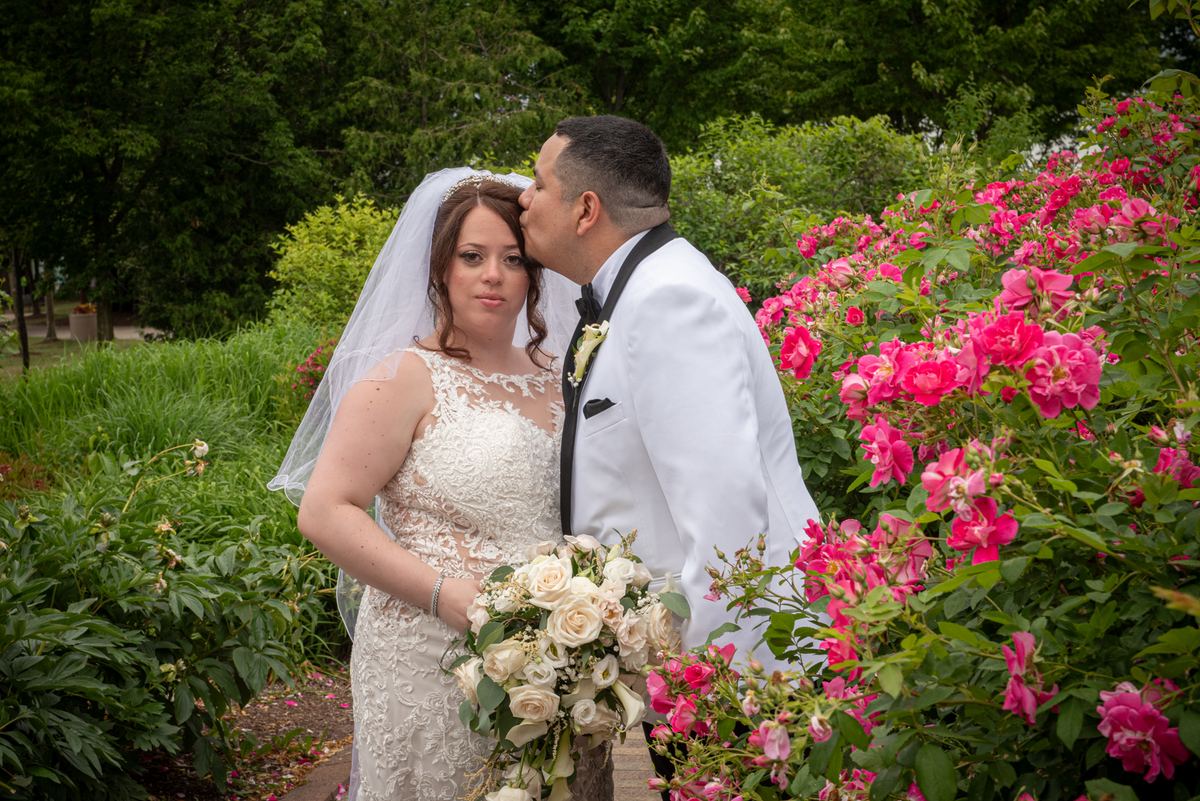 A bride in a lace gown and veil stands beside her groom as he kisses her cheek near pink roses in a garden.