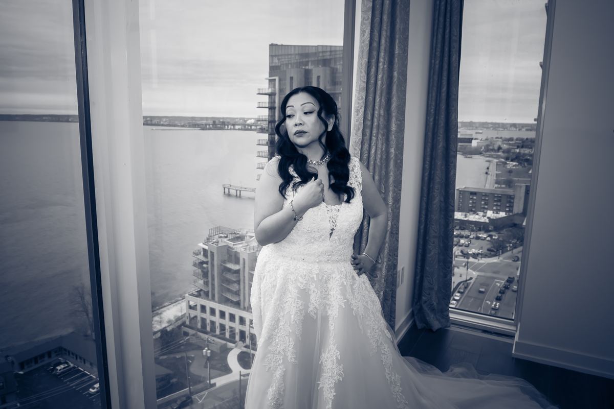 A bride in a lace gown stands by a window, gazing at a waterfront cityscape, her hand resting on her chest.