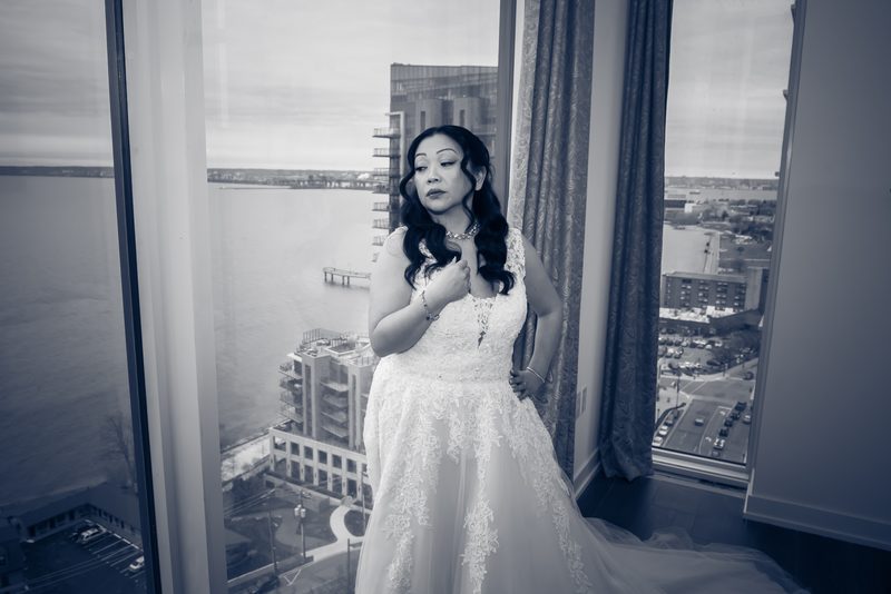 A bride in a lace gown stands by a window, gazing at a waterfront cityscape, her hand resting on her chest.