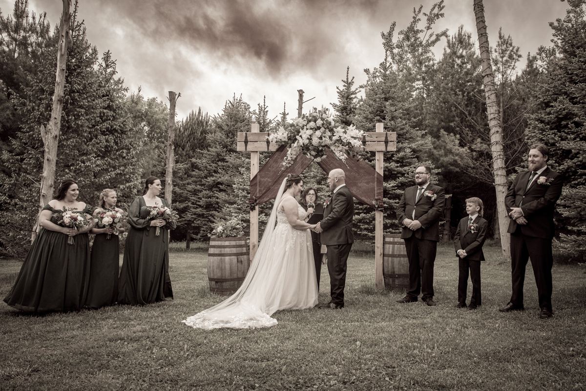 Bride and groom exchange vows under a rustic wooden arch adorned with flowers, surrounded by bridesmaids, groomsmen.