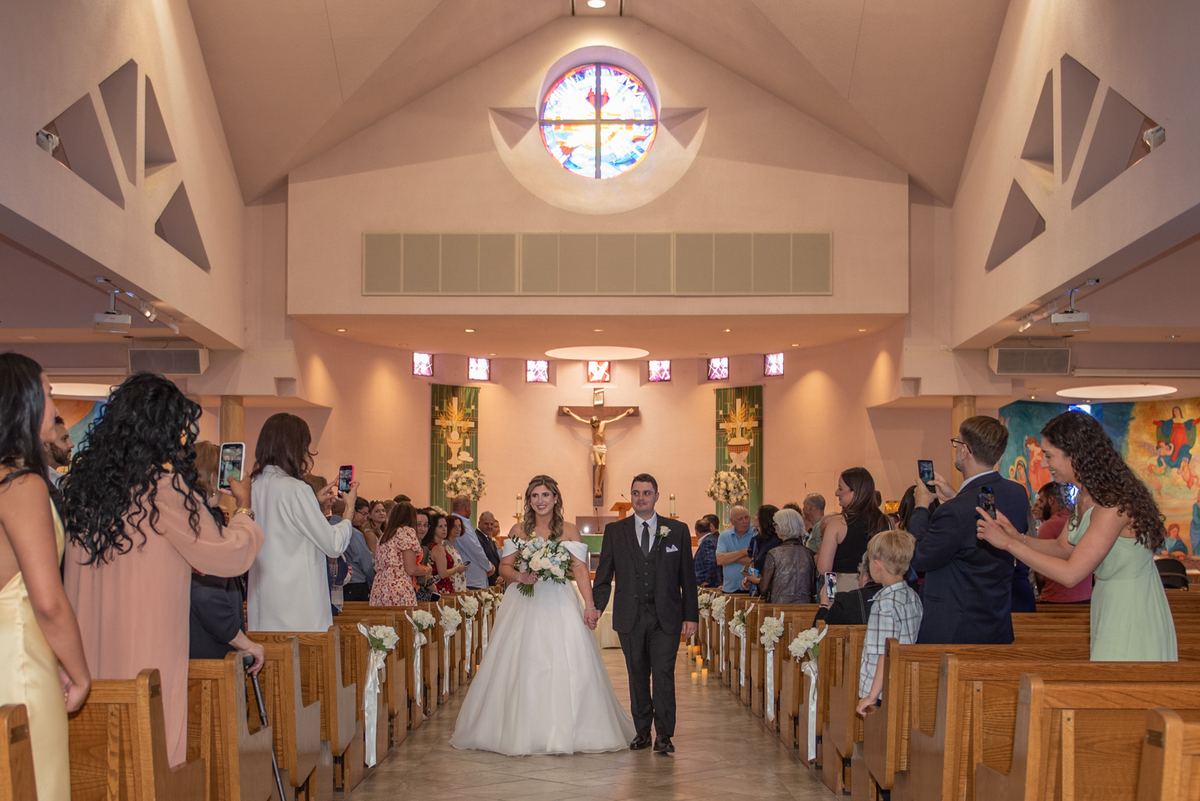 A bride and groom walk down the aisle of a modern church, illuminated by warm light from a stained-glass window.
