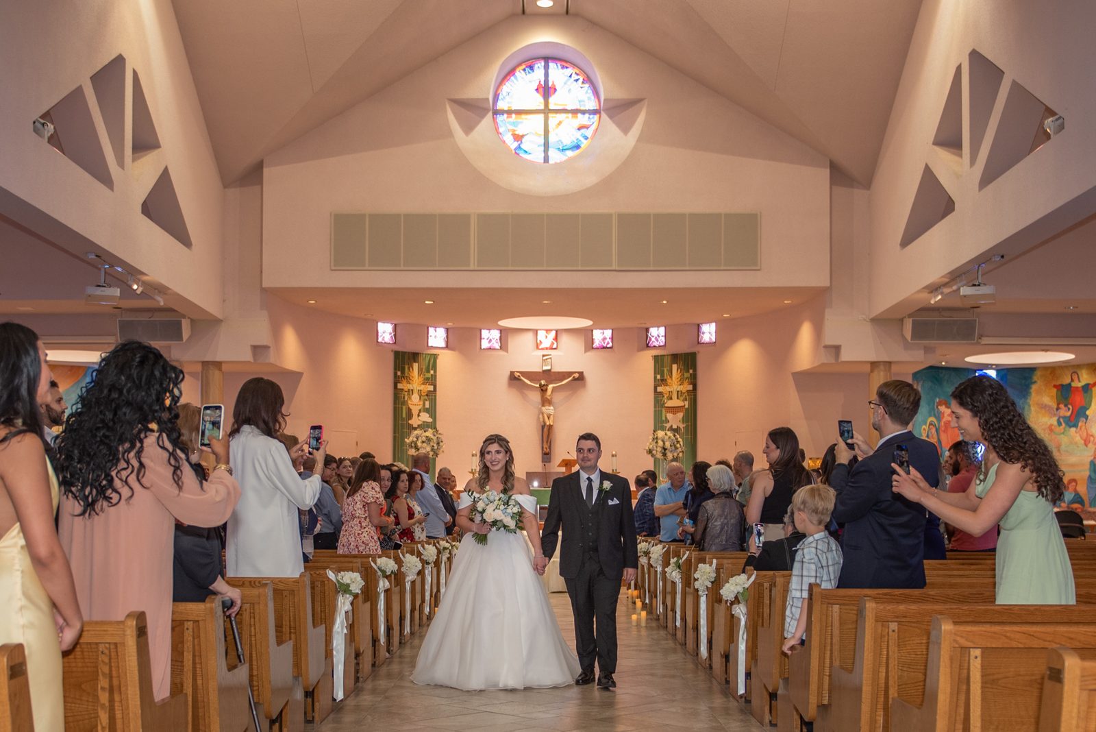 A bride and groom walk down the aisle of a modern church, illuminated by warm light from a stained-glass window.