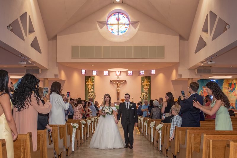 A bride and groom walk down the aisle of a modern church, illuminated by warm light from a stained-glass window.