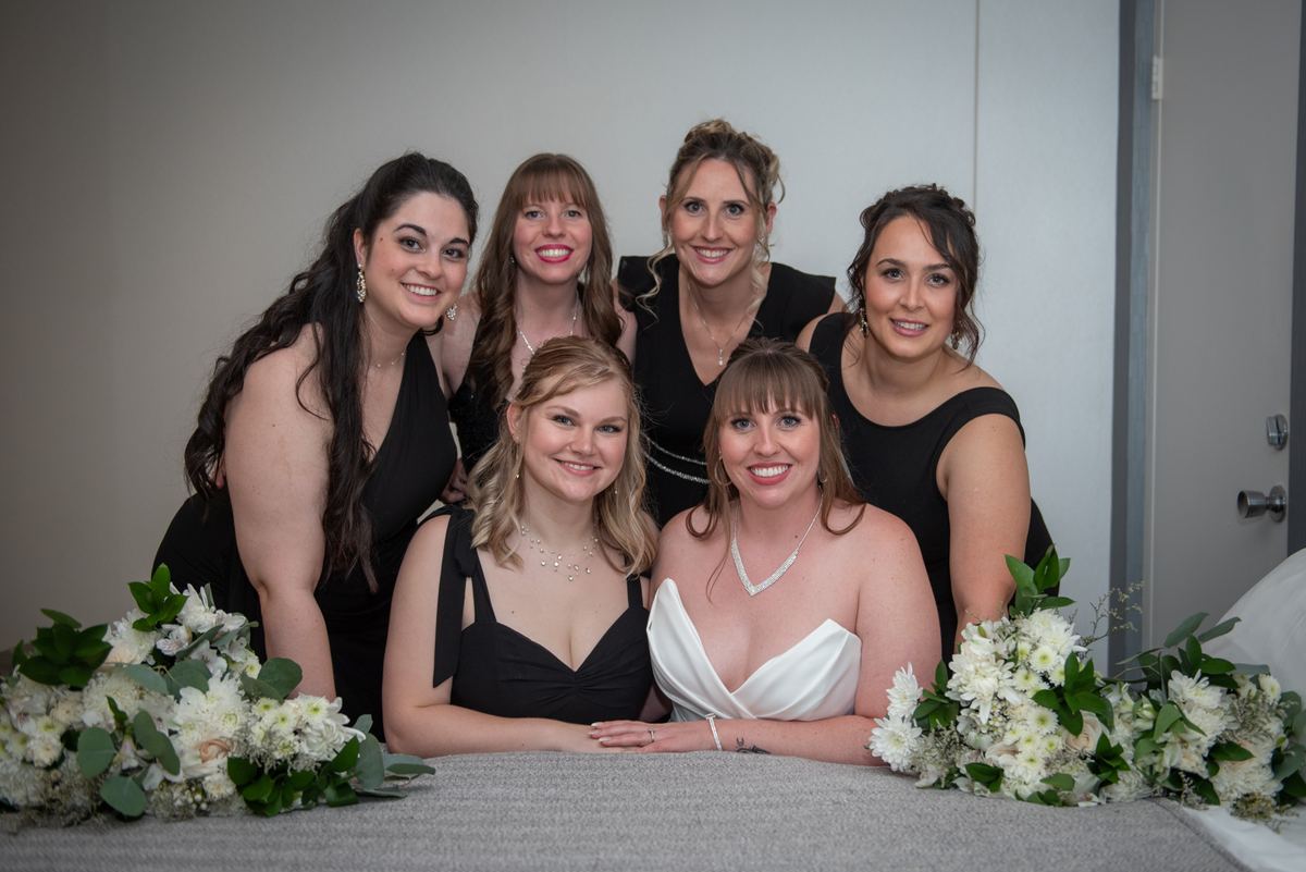 Bride in white gown surrounded by smiling bridesmaids in black dresses, all looking at camera near floral arrangements.