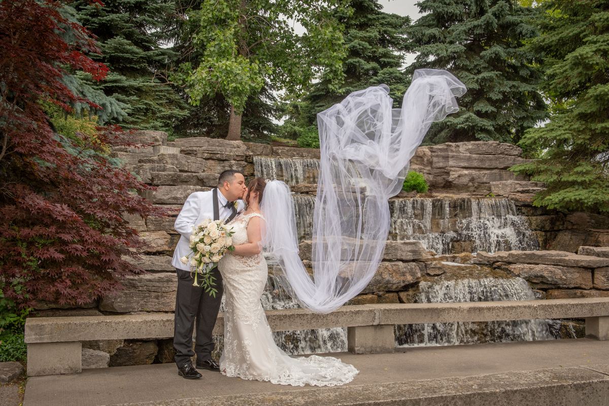 Lisa and Eduardo share a tender kiss beside a stone waterfall, with Lisa's veil billowing in the wind.
