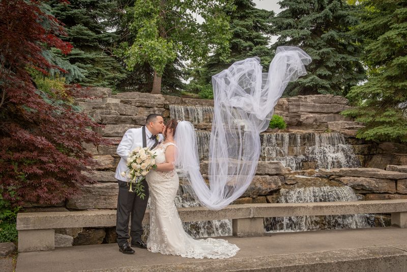 Lisa and Eduardo share a tender kiss beside a stone waterfall, with Lisa's veil billowing in the wind.