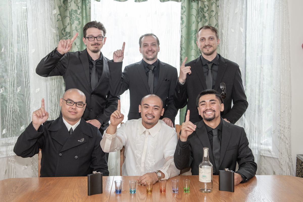 Six formally dressed men gather around a table, smiling and pointing upward in unison during a toast.