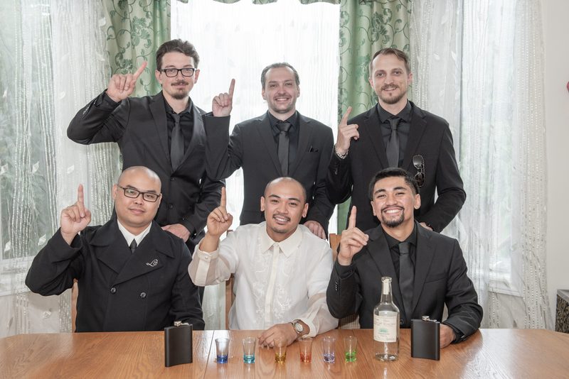 Six formally dressed men gather around a table, smiling and pointing upward in unison during a toast.