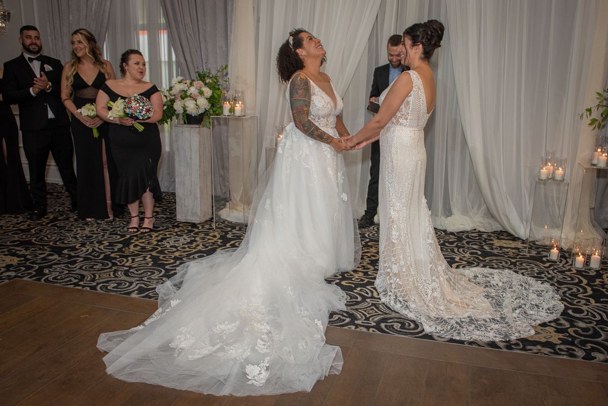 Two brides exchange vows in a warmly lit room, surrounded by applauding guests and floral arrangements.
