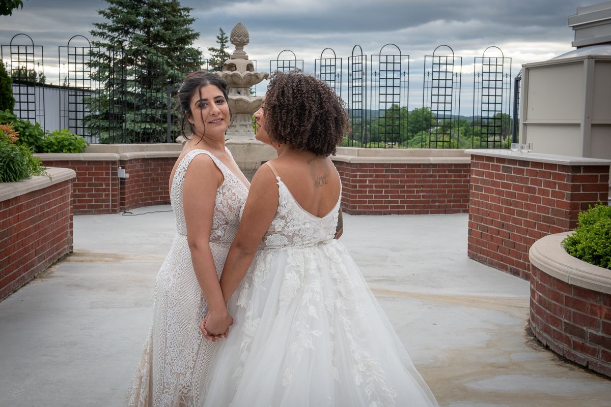 Two brides hold hands and smile on a rooftop terrace, surrounded by brick planters and ornamental fencing.