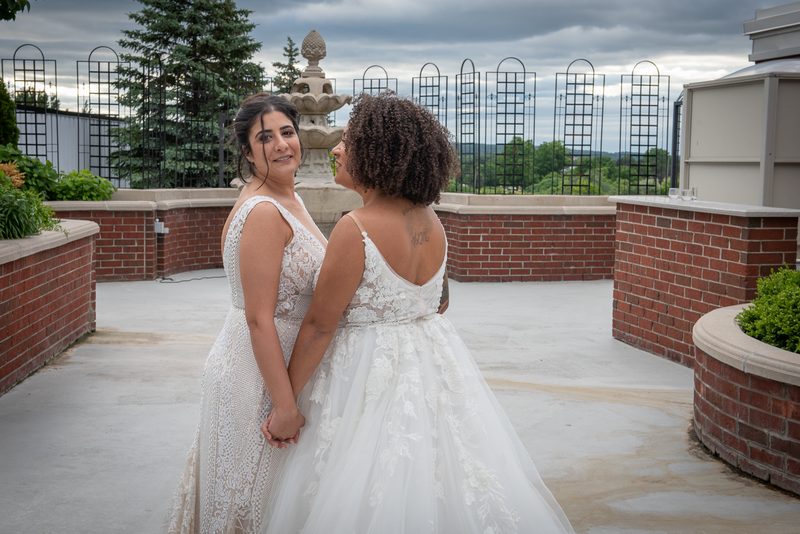 Two brides hold hands and smile on a rooftop terrace, surrounded by brick planters and ornamental fencing.