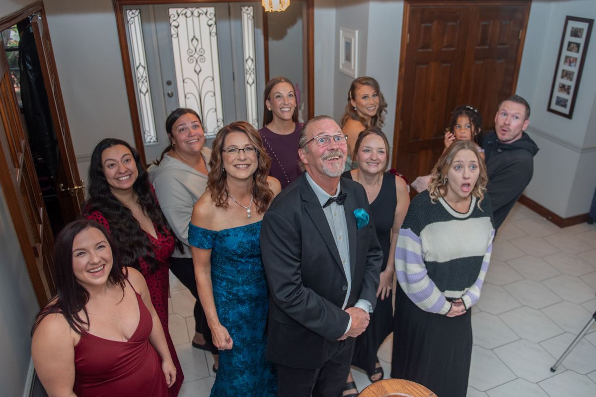 A group of family and friends in formal attire react with delight to an unexpected moment indoors, near a decorative door.