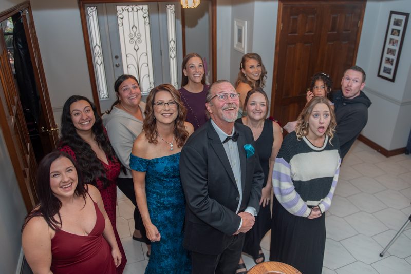A group of family and friends in formal attire react with delight to an unexpected moment indoors, near a decorative door.
