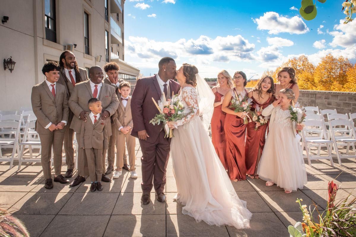 Bride and groom kiss joyfully outdoors, surrounded by smiling wedding party against autumn backdrop.