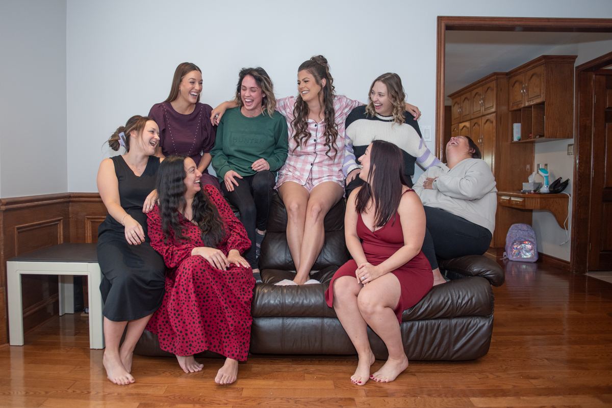 Seven women laughing and lounging barefoot on a leather couch in a cozy living room, daytime natural light.