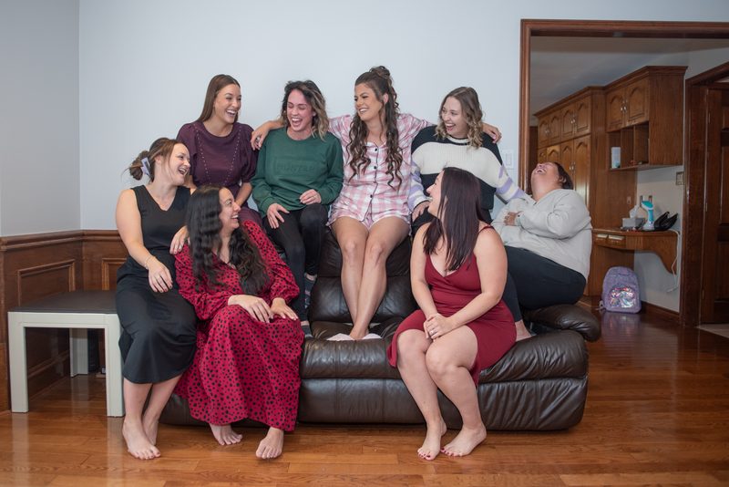 Seven women laughing and lounging barefoot on a leather couch in a cozy living room, daytime natural light.