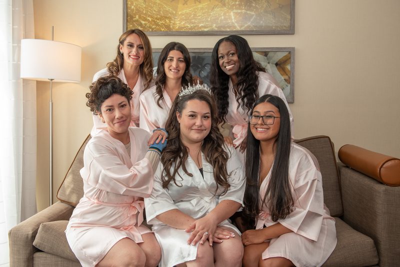 Bride in a tiara sits with smiling bridesmaids in satin robes, illuminated by warm light from a floor lamp.