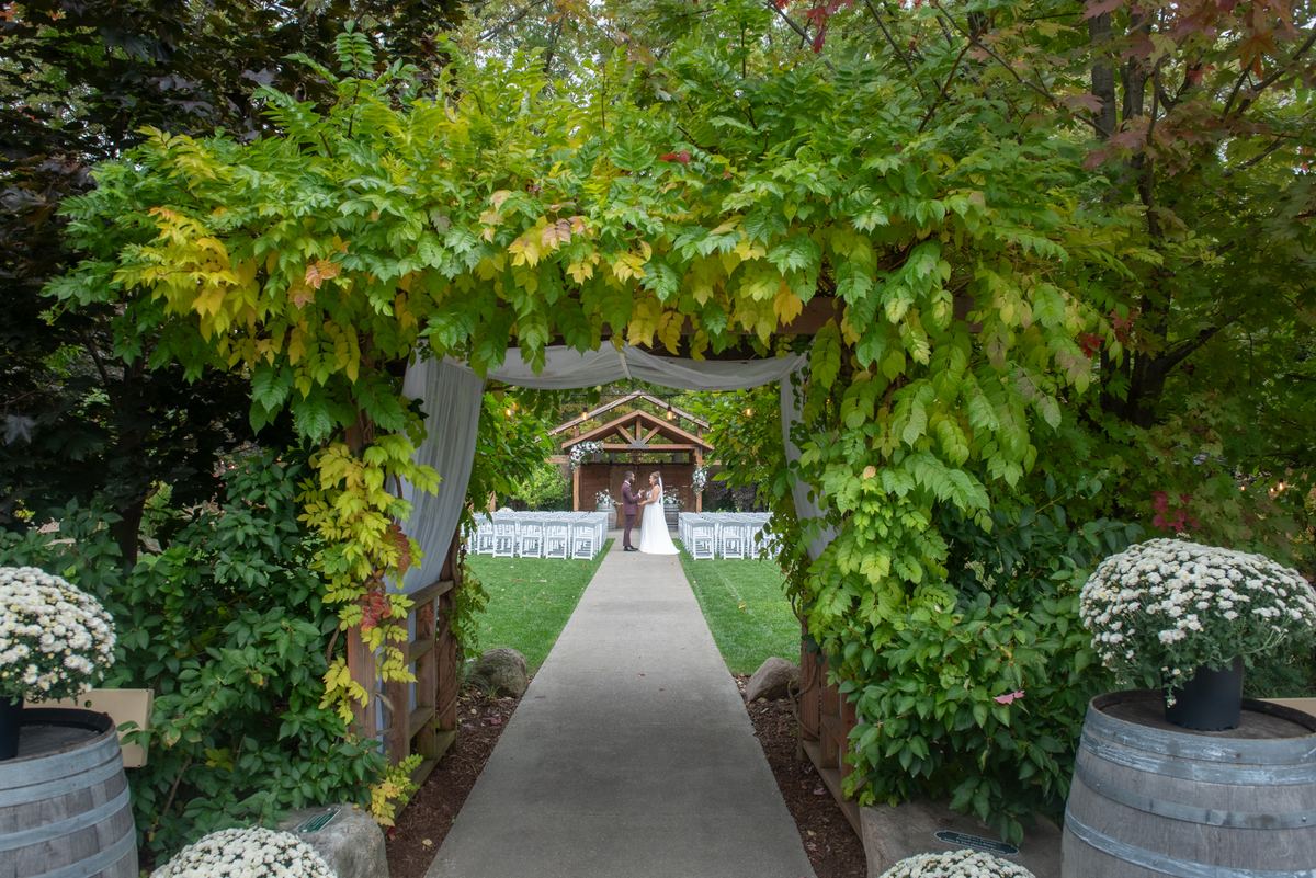 A bride and groom stand beneath a vine-covered arbor, surrounded by autumn foliage and chrysanthemums.