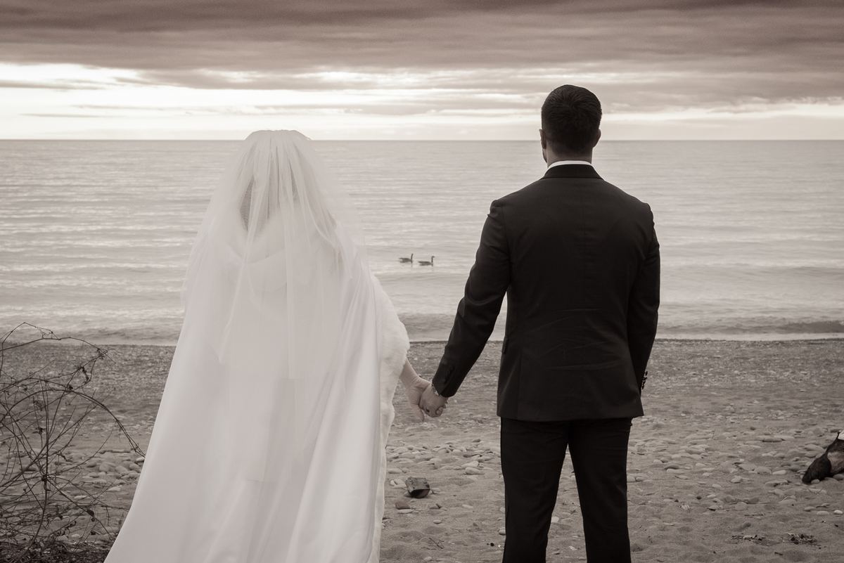 A newlywed couple stands on a pebbled beach, gazing at the sea under an overcast sky, hands clasped.