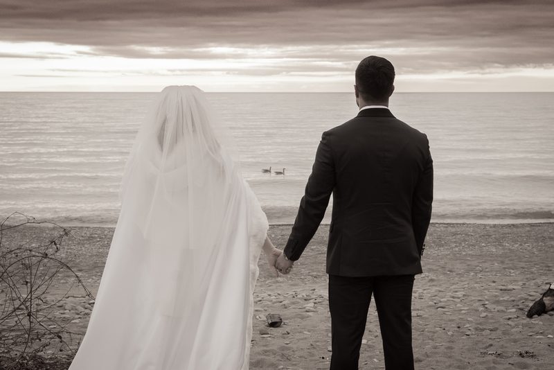 A newlywed couple stands on a pebbled beach, gazing at the sea under an overcast sky, hands clasped.