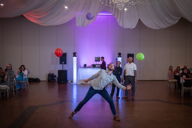 A man in a patterned shirt and jeans dances energetically as guests watch from chairs, surrounded by purple lighting.