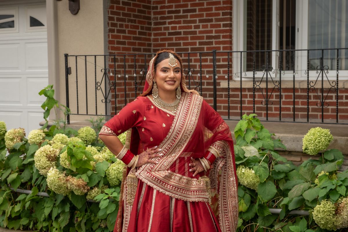 A bride in a red and gold lehenga with intricate embroidery stands confidently by a brick house surrounded by hydrangeas.