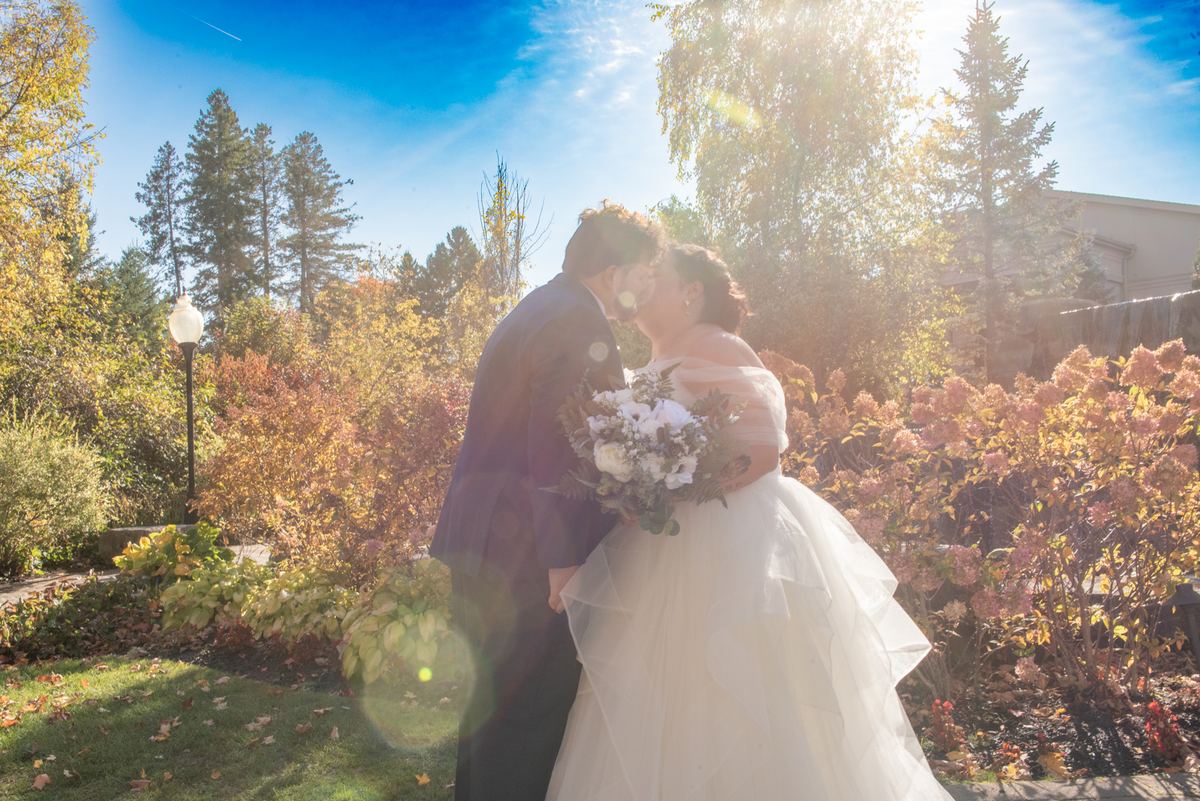 A bride and groom kiss in an autumn garden, surrounded by colorful leaves and lens flares from the sun.