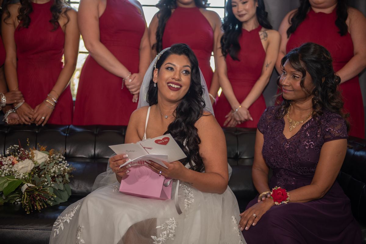 A bride smiles while holding a pink card, with bridesmaids in red dresses and a woman in purple beside her.