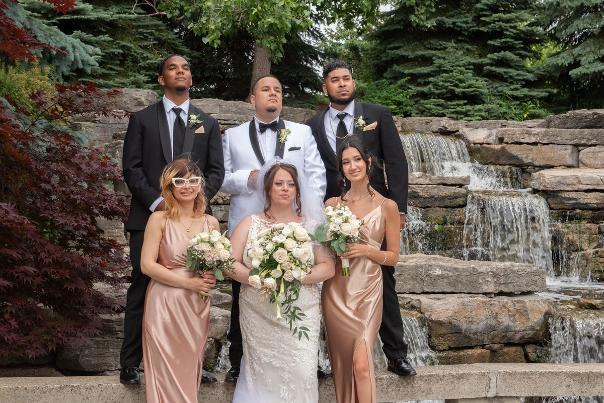 A bride in a white lace gown and veil stands beside a stone waterfall with bridesmaids and groomsmen in formal attire.