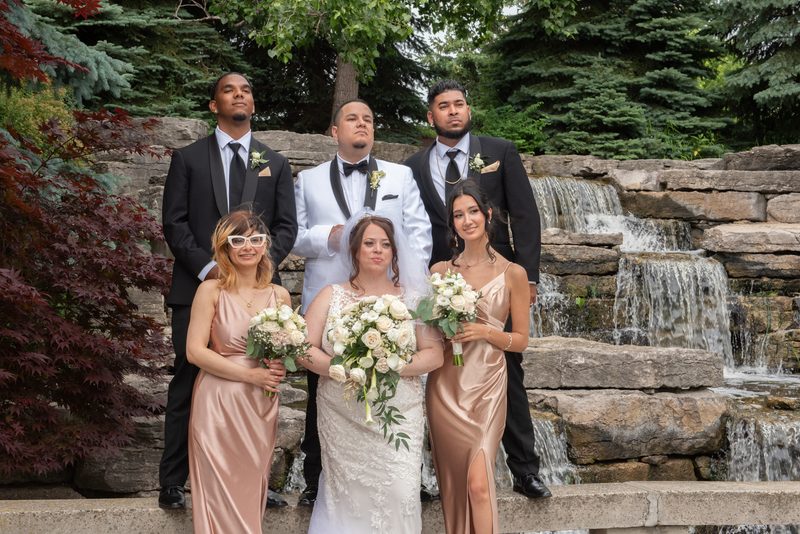 A bride in a white lace gown and veil stands beside a stone waterfall with bridesmaids and groomsmen in formal attire.