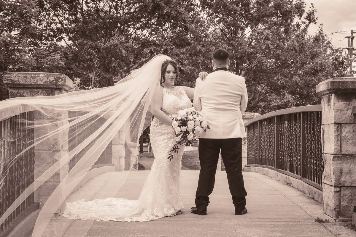 A bride in a lace gown and veil stands beside her groom on a stone bridge, placing her hand gently on his shoulder.