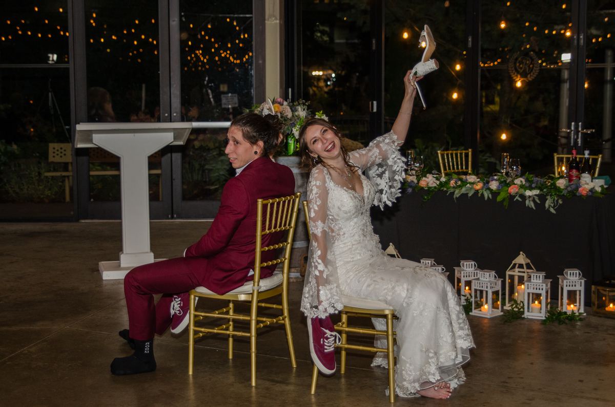 Bride and groom seated together, she holding up her heel playfully as he looks amused, surrounded by string lights.