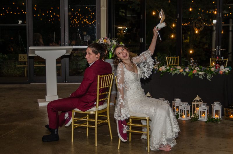 Bride and groom seated together, she holding up her heel playfully as he looks amused, surrounded by string lights.