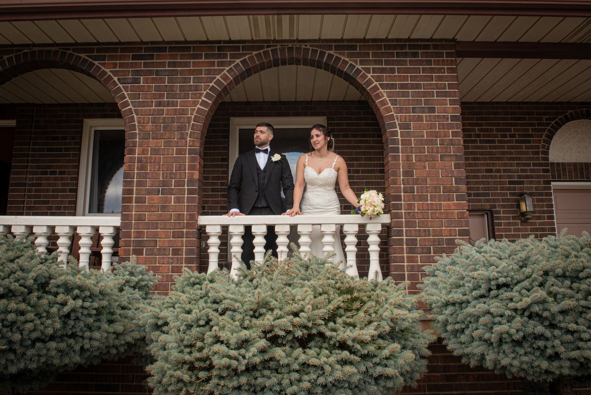 A bride and groom stand on a brick balcony, framed by arches and evergreen bushes, gazing toward the side in soft daylight.