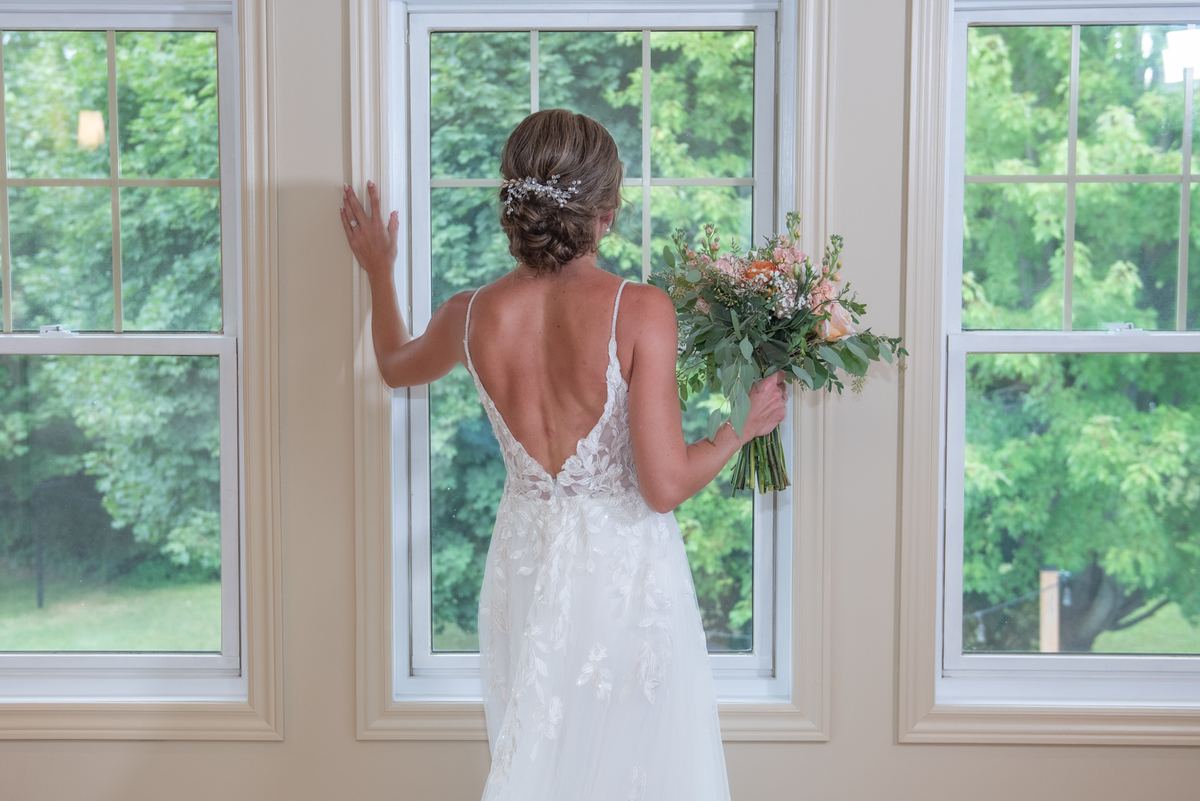 A bride in an elegant lace gown with a deep open back holds flowers and gazes out a window framed by greenery.