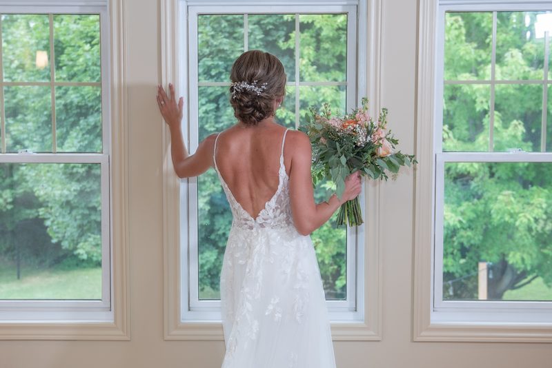 A bride in an elegant lace gown with a deep open back holds flowers and gazes out a window framed by greenery.