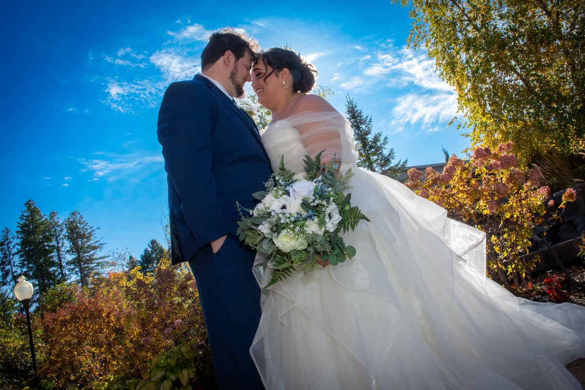 A bride and groom share an intimate moment outdoors under a blue sky with autumn foliage around them.